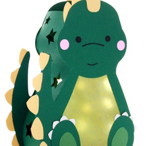 Pode incluir: Uma lanterna de papel verde em forma de dinossauro com um chifre amarelo, espinhos amarelos na cauda e detalhes brancos ao redor dos p&eacute;s. A lanterna tem um interior amarelo claro e estrelas recortadas nas laterais.