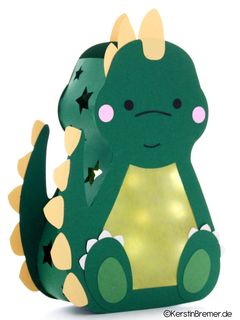 Pode incluir: Uma lanterna de papel verde em forma de dinossauro com um chifre amarelo, espinhos amarelos na cauda e detalhes brancos ao redor dos p&eacute;s. A lanterna tem um interior amarelo claro e estrelas recortadas nas laterais.
