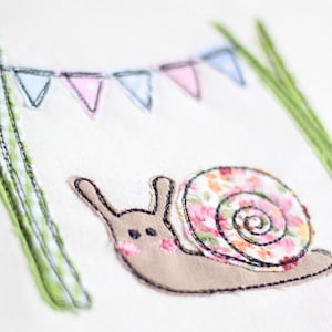Puede incluir: Un caracol marrón con una concha floral rosa y blanca está bordado sobre un fondo de tela blanca. Dos tallos verdes con patrones de cuadros blancos y verdes están bordados a cada lado del caracol. Una hilera de banderas triangulares rosas y azules está bordada sobre el caracol.