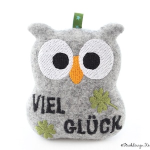 May include: A gray felt owl-shaped ornament with large white eyes, black pupils, and an orange beak. The German text "VIEL GLÜCK" is embroidered on the front, along with green shamrock accents. A green ribbon is attached at the top.