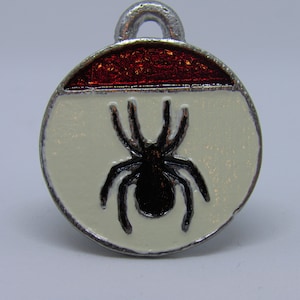 Może przedstawiać: Srebrny charm z czerwono-białym wzorem emaliowanym. Charm przedstawia czarnego pająka w środku.