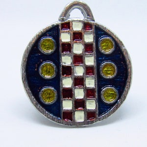 May include: A round, silver charm with a blue, red, and white checkered pattern and yellow circles. The charm has a loop at the top for attaching to a necklace or bracelet.