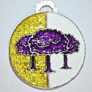 May include: A round pendant with a silver loop. The pendant is divided into two halves: one half is gold glitter, and the other is white. Three purple trees with white outlines are centered on the pendant.