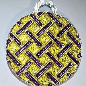 May include: A round, silver-toned pendant with a glittery yellow background and a woven pattern of purple rectangles. The pendant has a silver loop at the top for hanging.