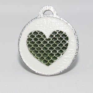 May include: A round, white pet tag with a silver-colored border and a loop for attachment. The tag features a green, scale-patterned heart in the center. The background is a plain white.