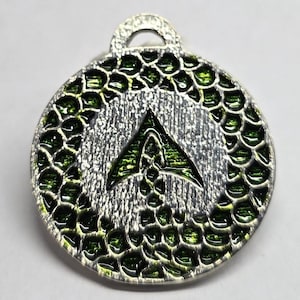 May include: Silver-toned pendant with a green and silver design. The pendant has a circular shape with a textured border of green scales. The center features a green arrow-like symbol on a textured silver background. A loop is at the top.