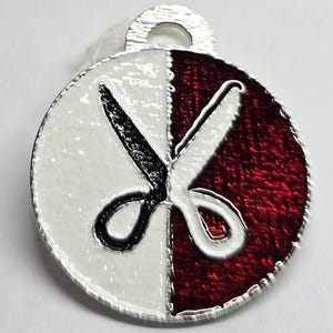 May include: A round, silver-toned charm with a split design. One half is white, the other is red. A black and white scissors graphic is overlaid on the charm. The charm has a small loop at the top for attachment.