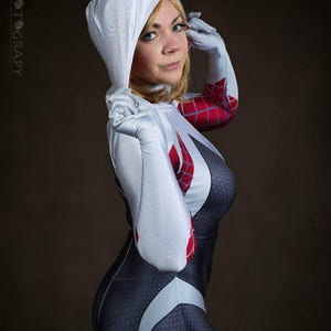 May include: A person in a Spider-Gwen costume. The costume is primarily black with a white spiderweb design and red accents. The hood and gloves are white. The background is a solid brown.