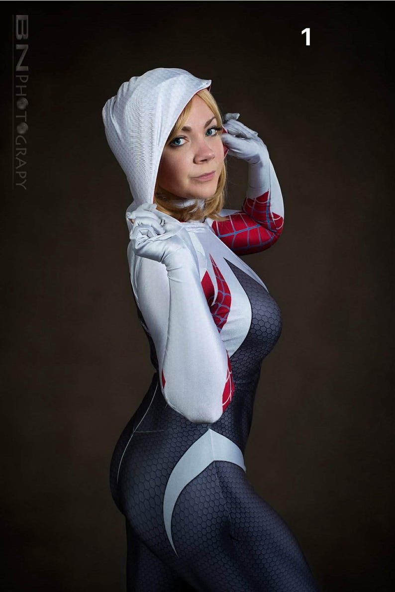 May include: A person in a Spider-Gwen costume. The costume is primarily black with a white spiderweb design and red accents. The hood and gloves are white. The background is a solid brown.