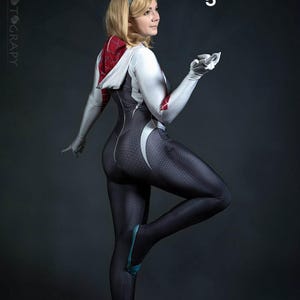 May include: A woman in a full-body cosplay suit, featuring a black and white design with a spiderweb pattern. The suit has a hood with red and white accents. The woman is wearing a black headband and gloves, with blue accents on the shoes. The number "5" is visible.