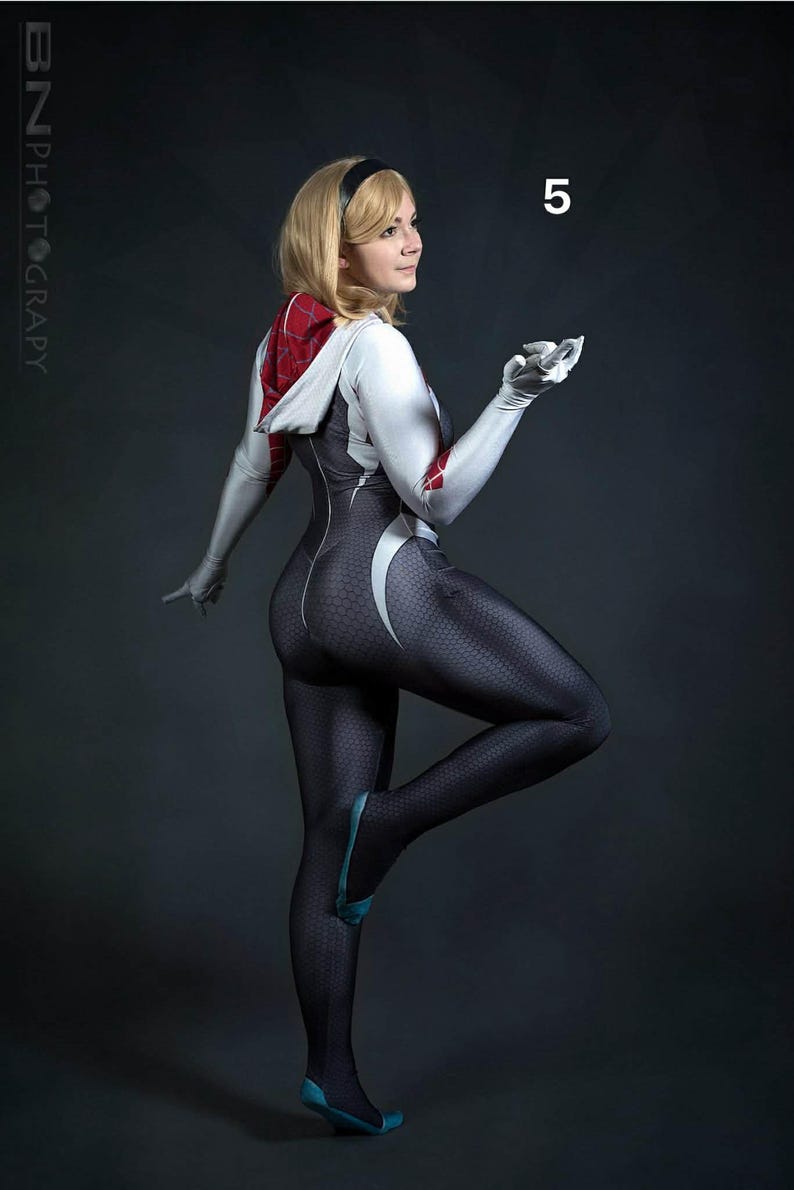 May include: A woman in a full-body cosplay suit, featuring a black and white design with a spiderweb pattern. The suit has a hood with red and white accents. The woman is wearing a black headband and gloves, with blue accents on the shoes. The number "5" is visible.