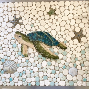 May include: A ceramic mosaic tile art piece depicting a sea turtle swimming in a sea of white and blue pebbles. The turtle is green and blue with a white shell. The pebbles are arranged in a random pattern.