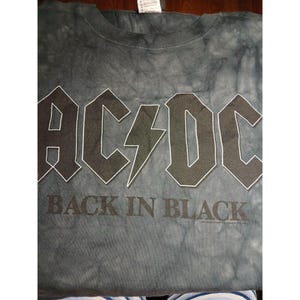 May include: A gray tie-dye t-shirt featuring the band name "AC/DC" in bold, white-outlined letters, with a lightning bolt graphic. The text "BACK IN BLACK" is printed below. The shirt is made of a soft material.