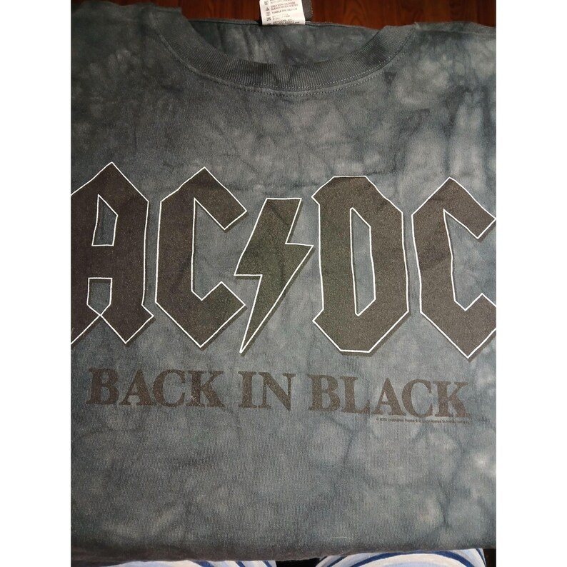 May include: A gray tie-dye t-shirt featuring the band name "AC/DC" in bold, white-outlined letters, with a lightning bolt graphic. The text "BACK IN BLACK" is printed below. The shirt is made of a soft material.