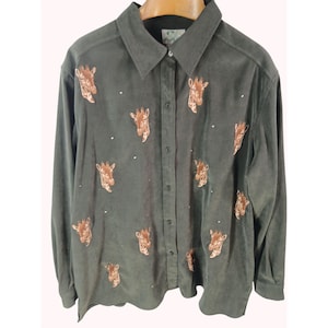 May include: A long-sleeved, button-down shirt in a muted green hue. The shirt features embroidered giraffe heads in shades of brown, scattered across the fabric, with small, sparkling embellishments. The collar is a classic point style.