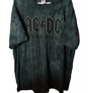 May include: Dark green tie-dye t-shirt featuring the band name "AC/DC" in white block letters with a lightning bolt in the center. Below the band name is the text "BACK IN BLACK". The shirt has short sleeves.