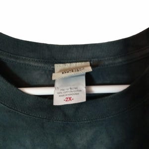 May include: Close-up of a dark green t-shirt collar with a tag. The tag reads "100% Cotton/Coton Made in Honduras" and "-2X-". The shirt is hanging on a white hanger.