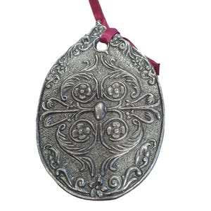 Può includere: Ornamento ovale color argento con un intricato design floreale e a volute. Un nastro bordeaux è attaccato in alto. L'ornamento presenta un motivo in rilievo dettagliato e un piccolo foro per appenderlo.