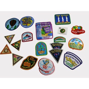 Puede incluir: Una colección de parches de Girl Scout con varios diseños. Los parches incluyen imágenes de un globo aerostático, un árbol, un pájaro y texto como "Happy Birthday" y "GS Cookies 2000". Los parches son de varios colores, incluyendo azul, verde y naranja.