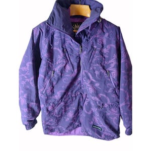 May include: A purple jacket with a floral pattern and a high collar. The jacket has a zipper closure, multiple pockets, and the word "HAVOC" printed on the bottom right. The jacket is hanging on a wooden hanger.