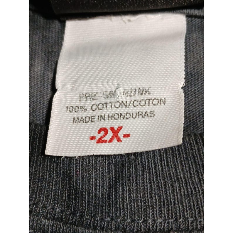 May include: Close-up of a white clothing tag with black text. The tag reads "PRE-SHRUNK 100% COTTON/COTON MADE IN HONDURAS" and has a red "-2X-" marking. The tag is attached to a dark gray fabric.