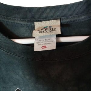 May include: Close-up of a dark green Liquid Blue t-shirt tag. The tag reads "LIQUID BLUE" and includes a size 2X label. The shirt is made of 100% cotton and made in Honduras.
