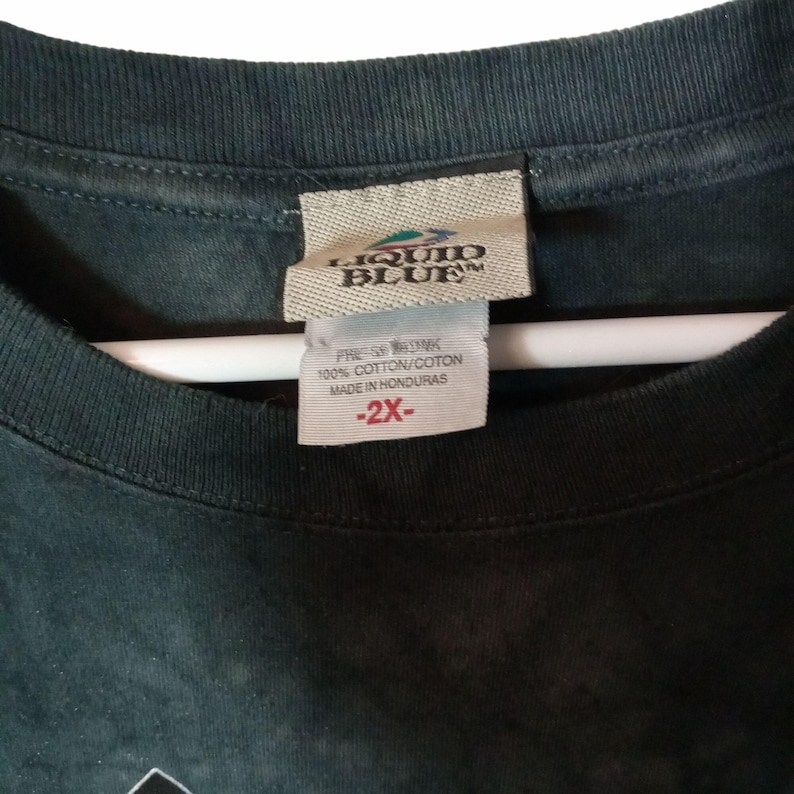 May include: Close-up of a dark green Liquid Blue t-shirt tag. The tag reads "LIQUID BLUE" and includes a size 2X label. The shirt is made of 100% cotton and made in Honduras.
