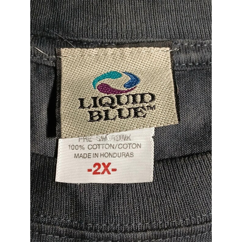 May include: Close-up of a gray t-shirt tag with the Liquid Blue logo in teal and blue. The tag also includes text that reads "100% Cotton/Coton Made in Honduras" and a size indicator of "-2X-".
