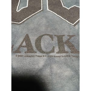 May include: Close-up of a blue-grey t-shirt with the word "ACK" in large, dark grey letters. The top portion of the letters "AC" are visible. The shirt has a textured appearance. Below the word "ACK" is text.