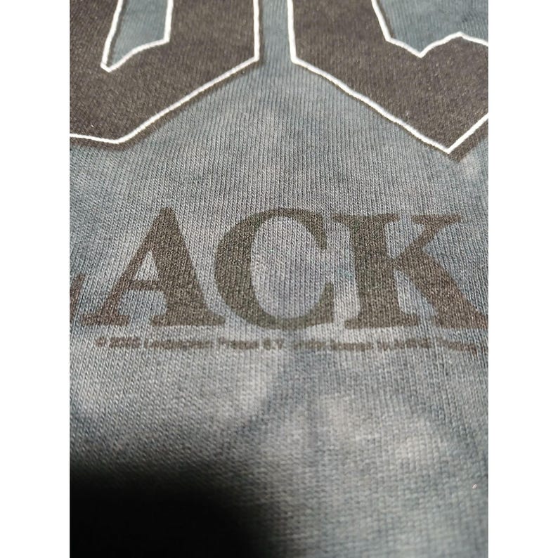 May include: Close-up of a blue-grey t-shirt with the word "ACK" in large, dark grey letters. The top portion of the letters "AC" are visible. The shirt has a textured appearance. Below the word "ACK" is text.