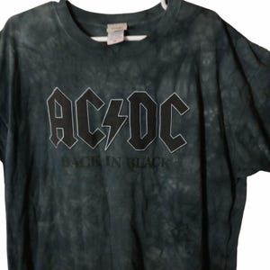 May include: Dark green tie-dye t-shirt featuring the band name "AC/DC" in a bold, outlined font with a lightning bolt graphic. Below the band name, the text "BACK IN BLACK" is printed in a simple font. The shirt is hanging on a white hanger.
