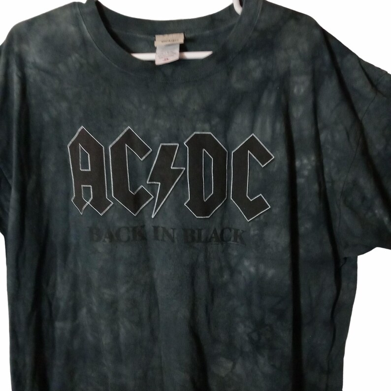May include: Dark green tie-dye t-shirt featuring the band name "AC/DC" in a bold, outlined font with a lightning bolt graphic. Below the band name, the text "BACK IN BLACK" is printed in a simple font. The shirt is hanging on a white hanger.