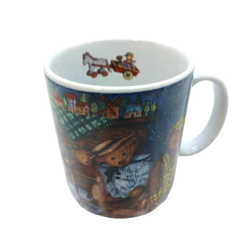 May include: A white ceramic mug featuring a dark blue exterior with a printed illustration of teddy bears, a windmill, and houses. The interior rim of the mug displays a small graphic of a horse pulling a wagon with a figure inside.