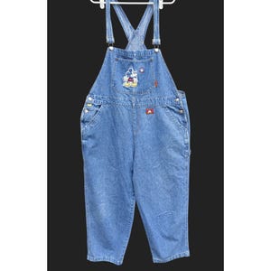 May include: Light blue denim overalls with adjustable straps and a bib pocket featuring a Mickey Mouse patch. The overalls have side pockets, a small red tag, and a relaxed fit. The garment is hanging on a white hanger.