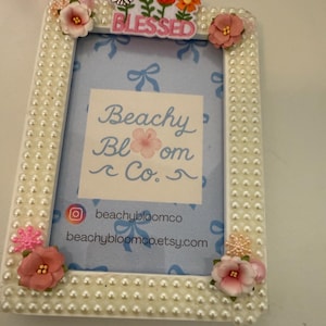 May include: A white rectangular photo frame, adorned with faux pearls and floral embellishments. The frame features a light blue backdrop with the text "Beachy Bloom Co." and the word "Blessed".
