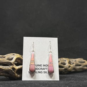 May include: A pair of dangle earrings with a geometric design. The earrings feature a pink stone inlay with silver-colored metal accents. The earrings are displayed on a white card with text that reads "GENUINE INDIAN ID CRAFT STERLING SIL".
