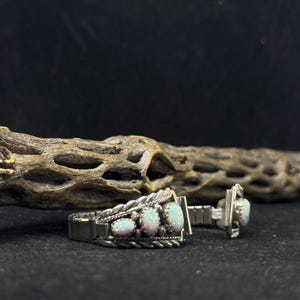 May include: Silver-toned cufflink set featuring three oval opals on each cufflink. The opals have a pearlescent sheen. The cufflinks have a braided rope design around the opals. The background is black.