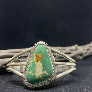 May include: A silver cuff bracelet with a green turquoise stone set in a silver bezel. The stone has a unique pattern of brown and white veins.