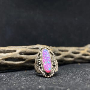 May include: A silver ring with an oval, pink opal center stone. The opal has a vibrant color with hints of blue and green. The ring has a detailed, ornate design around the stone and band.