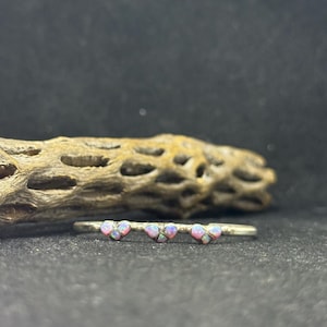 May include: A silver ring with three heart-shaped opal stones. The opals have a mix of pink, purple, and white colors. The ring is set against a dark background with a piece of dried wood.