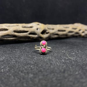 May include: A silver ring featuring two round, bright pink gemstones. The gemstones are set in a decorative, twisted metal setting. The ring is displayed on a dark gray surface.