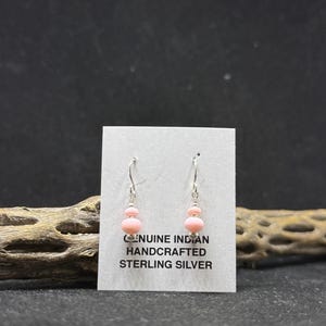 May include: Pink beaded earrings with sterling silver hooks. The earrings have a two-bead design, with a larger, oval-shaped bead at the bottom and a smaller, round bead at the top. The text on the card reads "GENUINE INDIAN HANDCRAFTED STERLING SILVER."