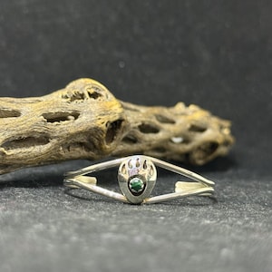 May include: A silver-toned bracelet featuring a bear paw design with a small, round, green gemstone at its center. The bracelet has a delicate, open design. The background includes a piece of dried wood.
