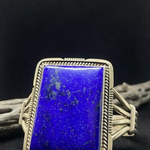 May include: A silver cuff bracelet featuring a large, rectangular lapis lazuli stone. The deep blue stone is set in a detailed silver frame with a twisted rope design. The bracelet has multiple silver bands on each side.
