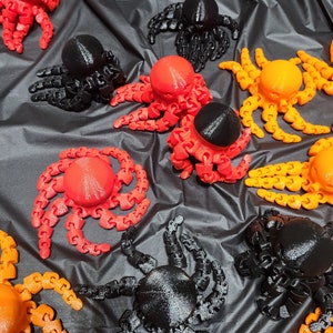 May include: A collection of 3D printed octopus figurines in various colors, including orange, red, and black. The octopus figurines have a segmented, articulated design, allowing them to be posed in different positions.