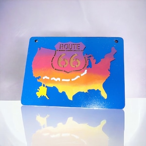 May include: A blue wooden sign with a cutout of the United States map. The map is painted in a gradient of pink, orange, and yellow. The sign also features a cutout of a Route 66 sign with the number 66 in white.