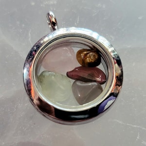 Puede incluir: Colgante de plata con un medallón de cristal transparente que contiene cuatro pequeñas piedras en forma de corazón: rosa, verde, marrón y roja.