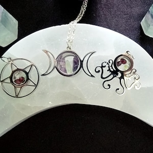 Peut inclure: Trois colliers en argent avec des pendentifs remplis de cristaux sur une plaque de recharge en sélénite en forme de croissant de lune blanc. Les pendentifs sont un pentagramme, une triple lune et un arbre de vie.