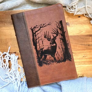 May include: A brown leather journal with a deer silhouette design. The journal has a two-tone design with a darker brown spine and a lighter brown front cover. The deer is depicted in a forest setting.