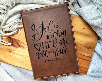 Könnte beinhalten: Ein braunes Lederjournal mit den Worten "Gott ist in ihr, sie wird nicht fallen" und dem Bibelvers "Psalm 46:5" in Schwarz eingraviert.